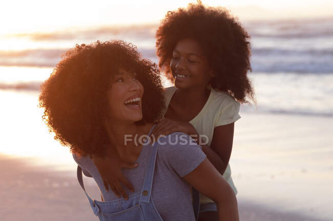 Smiling african american mother carrying her daughter piggyback at the beach smiling. healthy outdoor leisure time by the sea. — Fotografia de Stock