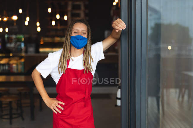 Портрет мужчины-баристы смешанной расы с дредами в маске для лица, склоняющейся в дверях кафе. независимый малый бизнес во время пандемии коронавируса. — стоковое фото