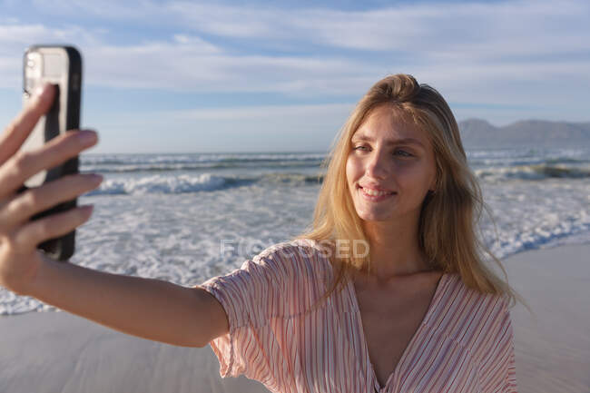 Белая женщина в пляжном покрытии делает селфи со смартфоном на пляже. здоровый отдых на открытом воздухе у моря. — стоковое фото