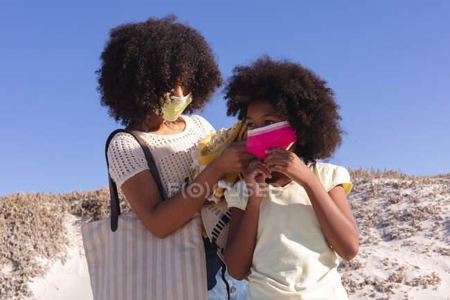 African american mother with daughter putting face mask on at the beach. family outdoor leisure time by the sea during coronavirus covid 19 pandemic. - foto de stock