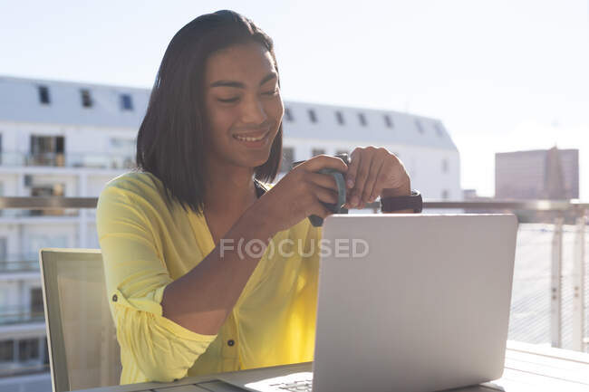 Усміхнена змішана трансгендерна жінка сидить за столом на сонячній терасі, використовуючи ноутбук, що тримає каву. перебування вдома в ізоляції під час карантину . — стокове фото