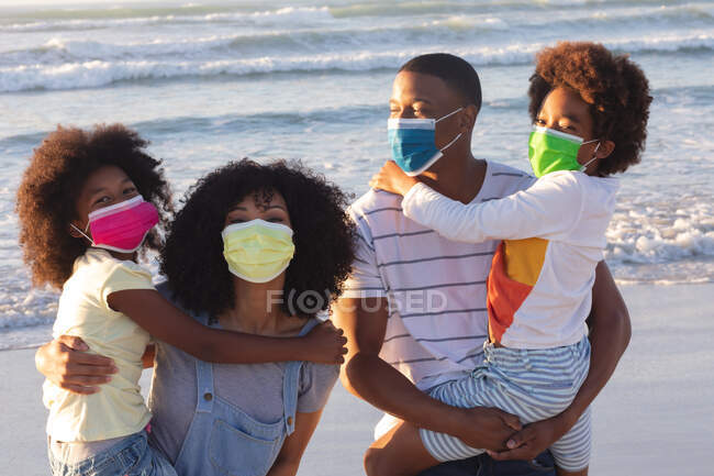 Портрет батьків-афроамериканців, які носять своїх двох дітей, одягнені в маски на пляжі. Час на відкритому повітрі біля моря під час пандемії коронавірусу 19. — стокове фото