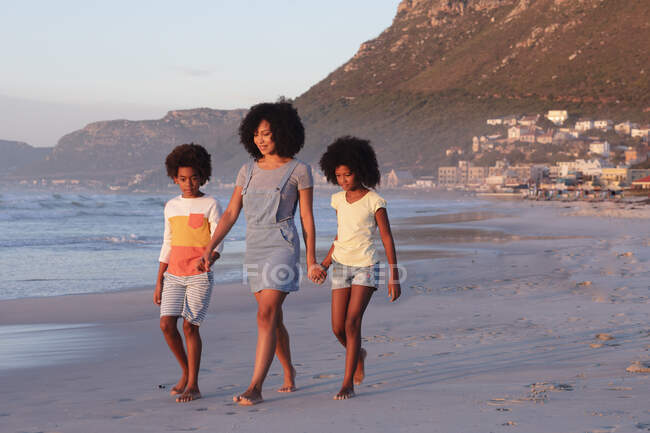 Африканская американская мать и двое детей ходят и держатся за руки на пляже. здоровый отдых на открытом воздухе у моря. — стоковое фото