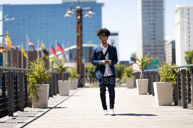 Hombre de raza mixta inteligentemente vestido con bigote caminando por la calle usando un teléfono inteligente. nómada digital, fuera y alrededor de la ciudad. - foto de stock