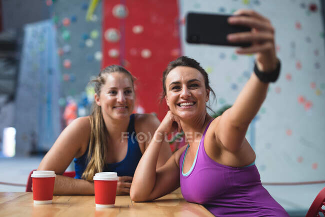 Dos mujeres caucásicas felices tomando selfie con teléfono inteligente en la cafetería en la pared de escalada interior. fitness y tiempo libre en el gimnasio. - foto de stock