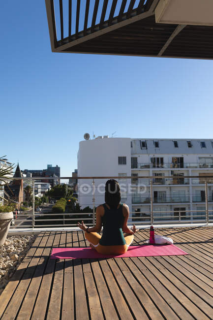 Visão traseira da mulher transgênero de raça mista praticando meditação de ioga no terraço do telhado ao sol. ficar em casa em isolamento durante o confinamento de quarentena. — Fotografia de Stock