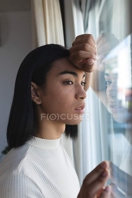 Mulher transgênero de raça mista olhando pela janela em pensamento no dia ensolarado. ficar em casa em isolamento durante o confinamento de quarentena. — Fotografia de Stock