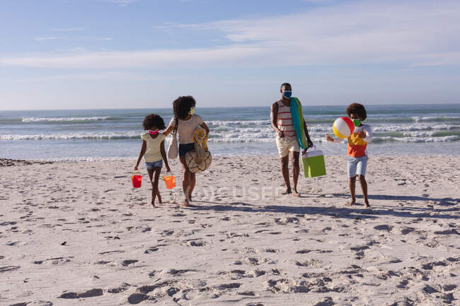 Pais afro-americanos e duas crianças segurando acessórios de praia andando na praia. família tempo de lazer ao ar livre junto ao mar durante coronavírus covid 19 pandemia. — Fotografia de Stock