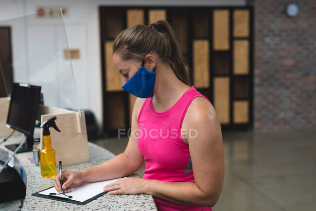 Mujer caucásica con máscara firmando documento en el mostrador del gimnasio. fitness y tiempo libre en el gimnasio durante coronavirus covid 19 pandemia. - foto de stock
