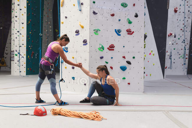 Zwei kaukasische Frauen an der Indoor-Kletterwand helfen beim Aufstehen. Fitness und Freizeit. — Stockfoto