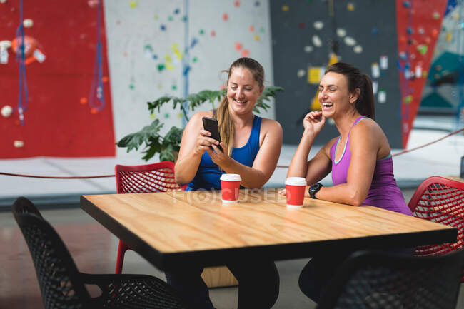 Dos mujeres caucásicas felices usando teléfono inteligente en la pared de escalada interior. fitness y tiempo libre en el gimnasio. - foto de stock