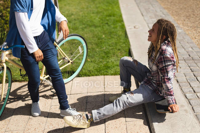 Deux heureux amis masculins métis assis sur le skateboard et le vélo dans la rue et parlant. mode de vie urbain vert, dans la ville. — Photo de stock