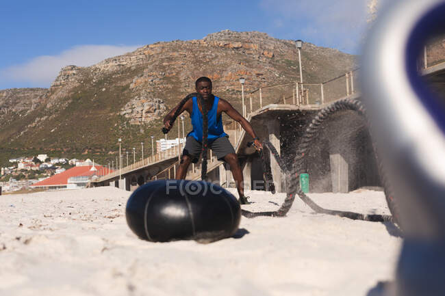 African american man exercising with rope on beach on sunny day. healthy outdoor lifestyle fitness training. — Stock Photo