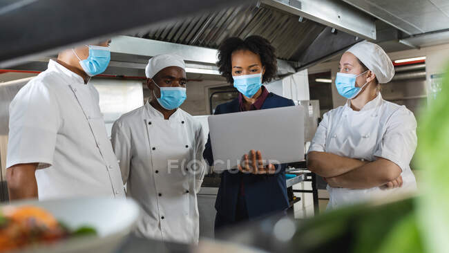 Diverso grupo de chefs profesionales que se reúnen con el gerente de cocina con máscaras faciales. trabajando en una ajetreada cocina de restaurante durante coronavirus covid 19 pandemia. - foto de stock