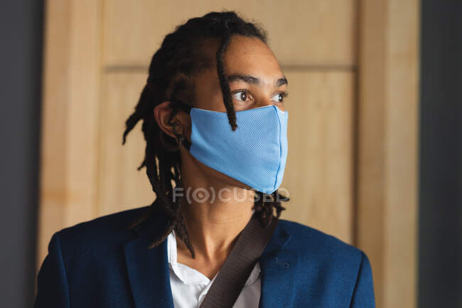 Портрет мужчины смешанной расы в маске для лица, стоящего в вестибюле отеля с плечевой сумкой. отель для деловых поездок во время пандемии коронавируса. — стоковое фото