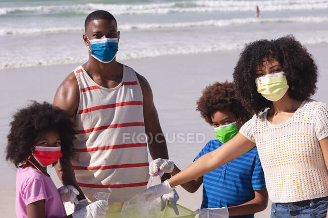 Батьки-афроамериканці з двома дітьми в масках на обличчі збирають сміття з пляжу. збереження пляжів родини під час коронавірусної ковини 19 пандемії.. — стокове фото