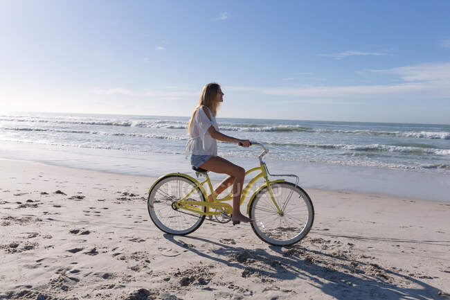 Mujer caucásica en bicicleta en la playa. tiempo de ocio al aire libre saludable junto al mar. - foto de stock