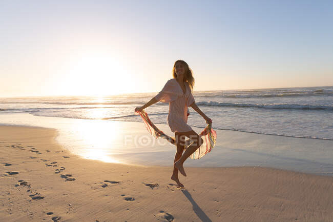 Hermosa mujer caucásica sosteniendo bufanda sonriendo mientras está de pie en la playa. verano playa vacaciones concepto. - foto de stock