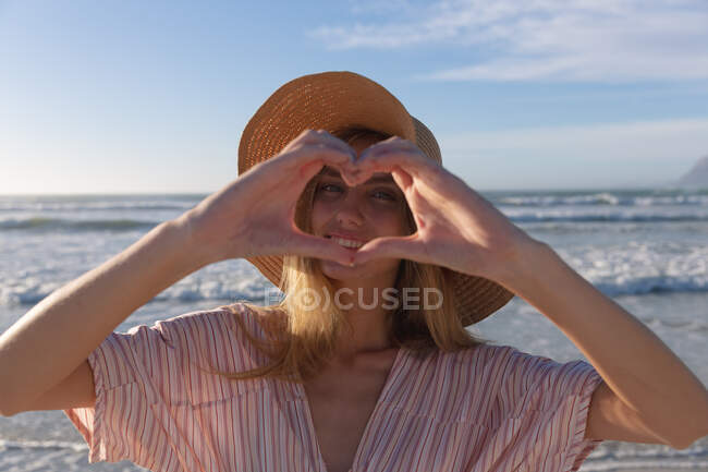 Caucasian woman wearing hat making heart shape looking at camera and smiling at the beach. healthy outdoor leisure time by the sea. — Stock Photo