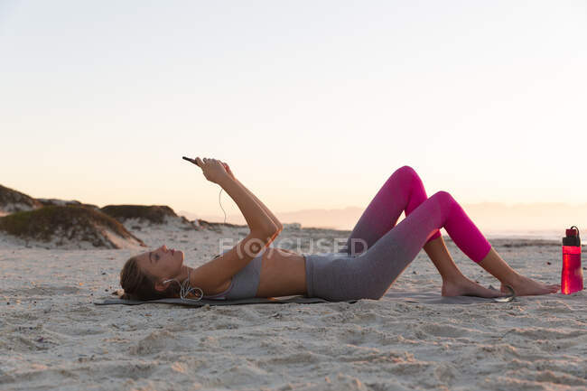 Mulher caucasiana se exercitando deitada na praia usando fones de ouvido usando smartphone. saúde e bem-estar, relaxando na praia ao nascer do sol. — Fotografia de Stock