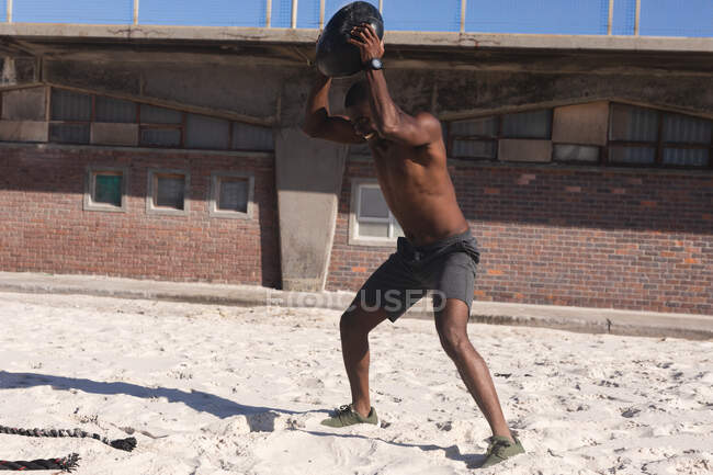 African american man exercising with weights on beach on sunny day. healthy outdoor lifestyle fitness training. — Foto stock