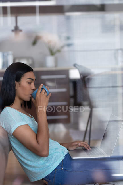 Женщина-трансгендер смешанной расы, работающая дома, пьет кофе на ноутбуке. оставаться дома в изоляции во время карантинной изоляции. — стоковое фото