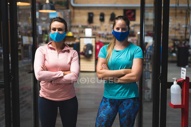 Retrato de dos mujeres caucásicas con máscaras de pie en el pasillo del gimnasio. fitness y tiempo libre en el gimnasio durante coronavirus covid 19 pandemia. - foto de stock