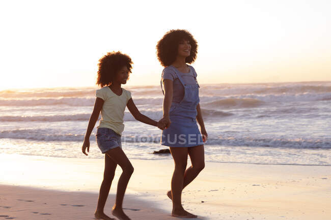 Улыбающиеся африканские американские мать и дочь, идущие и держащиеся за руки на пляже. здоровый отдых на открытом воздухе у моря. — стоковое фото