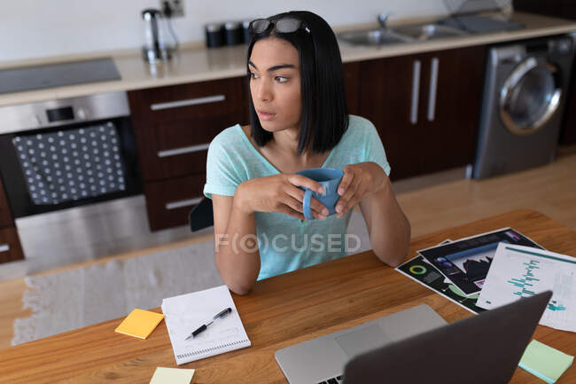 Mulher transgênero de raça mista trabalhando em casa usando laptop beber café. ficar em casa em isolamento durante o confinamento de quarentena. — Fotografia de Stock