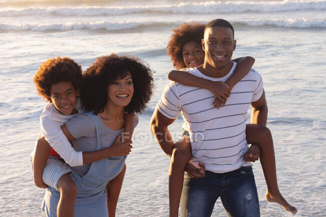 Smiling african american parents and carrying their two children piggyback at the beach. family outdoor leisure time by the sea. — Foto stock