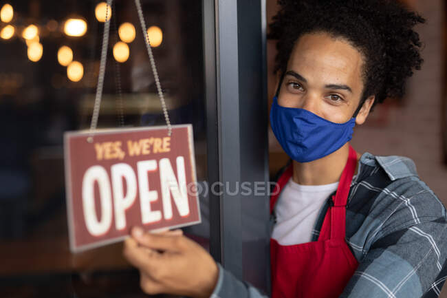 Portrait d'un barista masculin de race mixte portant un masque facial penché à la porte d'un café tenant un panneau ouvert. petite entreprise indépendante pendant une pandémie de coronavirus covid 19. — Photo de stock