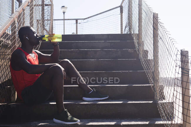 Afrikanisch-amerikanischer Mann, der an sonnigen Tagen Sport treibt, sich ausruht und Wasser auf Treppen trinkt. gesundes Outdoor-Fitness-Training. — Stockfoto