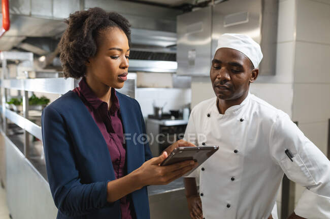 Carrera diversa gerente de cocina femenina discutiendo con chef profesional sobre la tableta. trabajando en una cocina ajetreada. - foto de stock