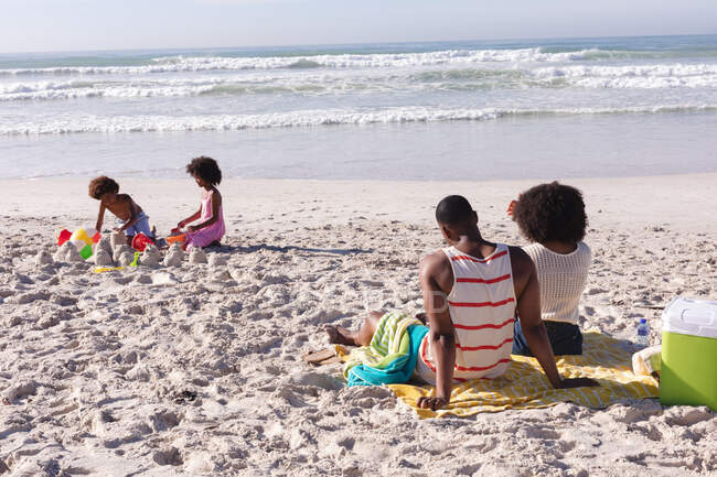 Afrikanisch-amerikanische Kinder beim Spielen mit Sand am Strand. mit Eltern auf dem Handtuch liegend. Familienfreizeit im Freien am Meer. — Stockfoto