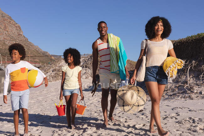 African american parents and two children holding beach accessories walking at the beach. family outdoor leisure time by the sea. — Foto stock