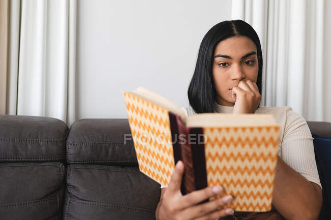 Mixed race transgender woman relaxing in living room sitting on couch reading book. staying at home in isolation during quarantine lockdown. — Stock Photo