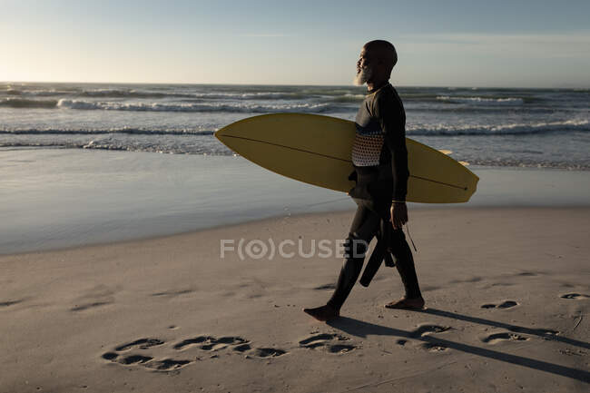 Hombre afroamericano mayor llevando tabla de surf caminando por la playa. viaje vacaciones retiro estilo de vida concepto - foto de stock