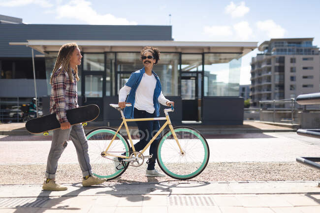 Two happy mixed race male friends wheeling bicycle carrying skateboard in the street and talking. green urban lifestyle, out and about in the city. — Stock Photo
