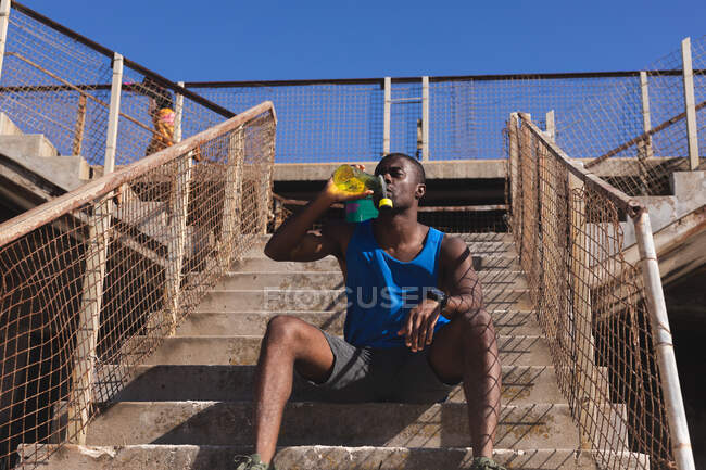 African american man exercising, resting, drinking water on stairs on sunny day. healthy outdoor lifestyle fitness training. — Photo de stock