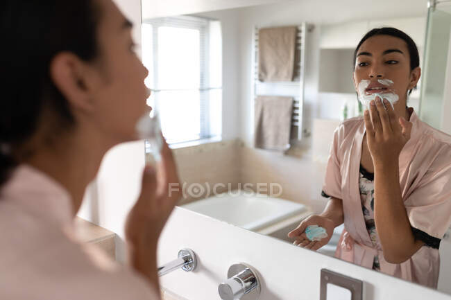 Mulher transexual de raça mista olhando no espelho do banheiro e colocando creme de barbear. ficar em casa em isolamento durante o confinamento de quarentena. — Fotografia de Stock