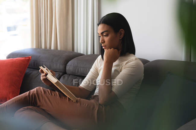 Mulher transexual de raça mista relaxante na sala de estar sentado no sofá livro de leitura. ficar em casa em isolamento durante o confinamento de quarentena. — Fotografia de Stock