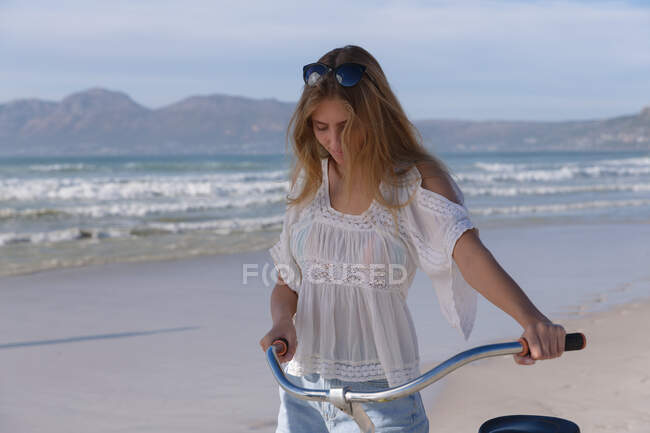 Кавказька жінка на білій верхівці і шорти, що ходять з велосипедом на пляжі. Здоровий вільний час на відкритому повітрі біля моря. — стокове фото