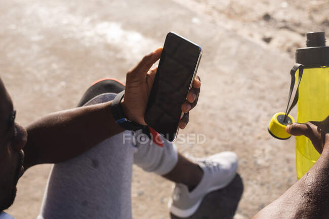 African american man exercising, resting, using smartphone on beach on sunny day. healthy outdoor lifestyle fitness training. — Stock Photo