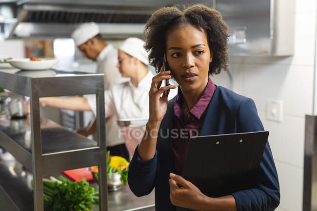Mixed race kitchen manager talking on the phone with professional chefs in background. working in a busy restaurant kitchen. — Stock Photo