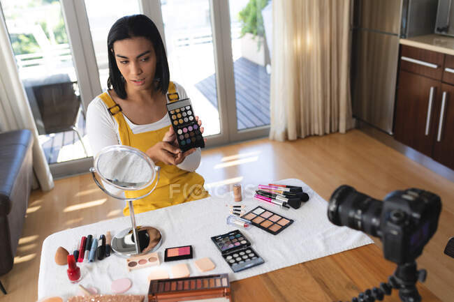Happy mixed race transgender woman making vlog using laptop and camera holding makeup and talking. staying at home in isolation during quarantine lockdown. — Stock Photo