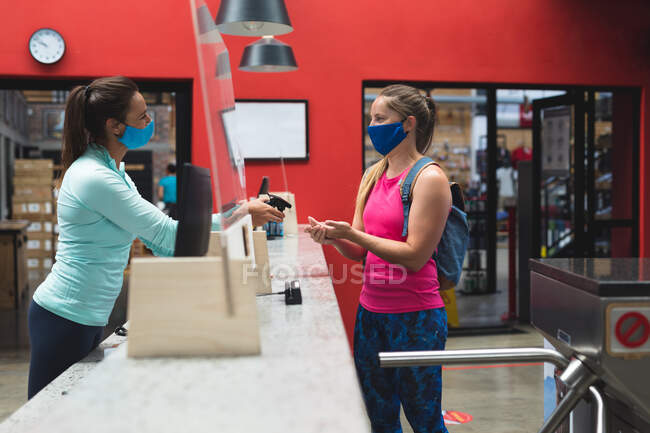 Empfangsdame und Kundin mit Masken desinfizieren im Fitnessstudio die Hände über dem Tresen. Fitness und Freizeit im Fitnessstudio während der Coronavirus-Pandemie 19. — Stockfoto