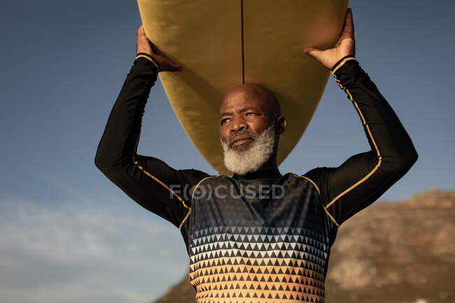 Ein älterer afrikanisch-amerikanischer Mann trägt am Strand ein Surfbrett auf dem Kopf. Reise Urlaub Ruhestand Lifestylekonzept — Stockfoto