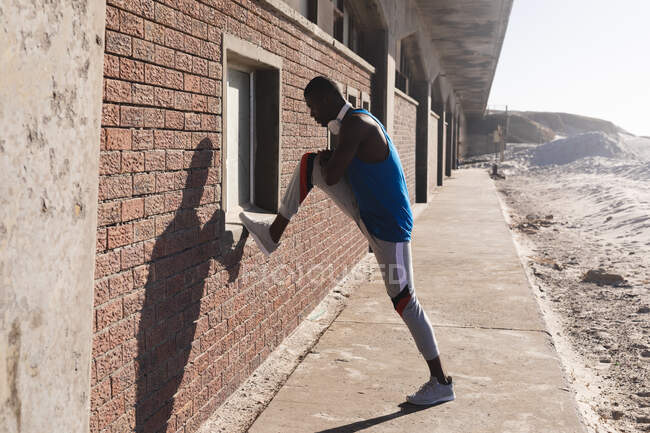 Afrikanisch-amerikanischer Mann, der im Freien trainiert, Kopfhörer trägt und sich an sonnigen Tagen dehnt. gesundes Outdoor-Fitness-Training. — Stockfoto