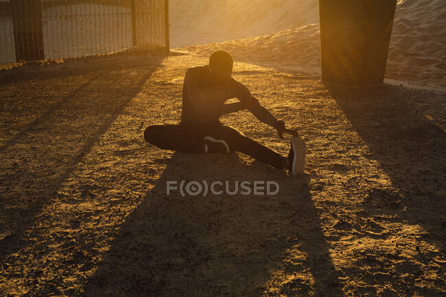 Homem afro-americano exercitando-se ao ar livre, estendendo-se sob ponte ao pôr do sol. treinamento de fitness ao ar livre saudável. — Fotografia de Stock