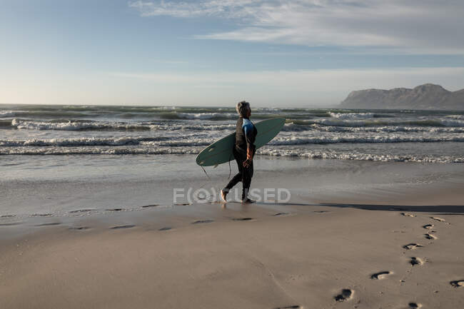 Donna afroamericana anziana che porta tavola da surf mentre cammina sulla spiaggia. viaggio vacanza pensionamento stile di vita — Foto stock
